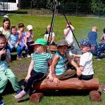 dzieci i panie siedzą na drewnianych ławkach wokół ogniska