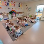 Dzieci siedzący w sali przedszkolnej na dywanie w trzech rzędach, jedno za drugim, mający rozłożone ręce na boki.