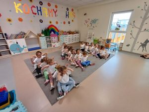 Dzieci siedzący w sali przedszkolnej na dywanie w trzech rzędach, jedno za drugim, mający rozłożone ręce na boki.