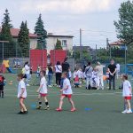 Dzieci biorące udział w konkurencjach sportowych na boisku. W tle stoją osoby dorosłe.