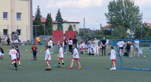 Dzieci biorące udział w konkurencjach sportowych na boisku. W tle stoją osoby dorosłe.