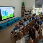 dzieci i dwie panie ubrane na niebiesko, siedzą na krzesełkach przed tv i oglądają bajkę edukacyjną