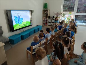 dzieci i dwie panie ubrane na niebiesko, siedzą na krzesełkach przed tv i oglądają bajkę edukacyjną