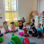Grupa dzieci siedzi kole razem z panią , która gra na gitarze. pośrodku koła stoi duża żaba , i zajączek.