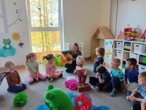 Grupa dzieci siedzi kole razem z panią , która gra na gitarze. pośrodku koła stoi duża żaba , i zajączek.