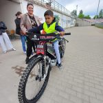 Chłopiec siedzący na motorze żużlowym. Z lewej strony stoją trzy Panie.