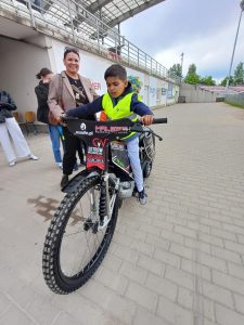 Chłopiec siedzący na motorze żużlowym. Z lewej strony stoją trzy Panie.