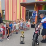 Grupa dzieci przed przedszkolem, razem z zaproszonym gościem. Dzieci oglądają żużlowy motor