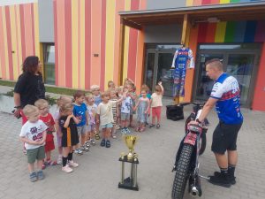 Grupa dzieci przed przedszkolem, razem z zaproszonym gościem. Dzieci oglądają żużlowy motor