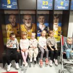 Sześcioro dzieci siedzących w szatni sportowców. Czworo siedzi na ławce, jeden chłopiec we wózku. W tle zdjęcia koszykarzy.