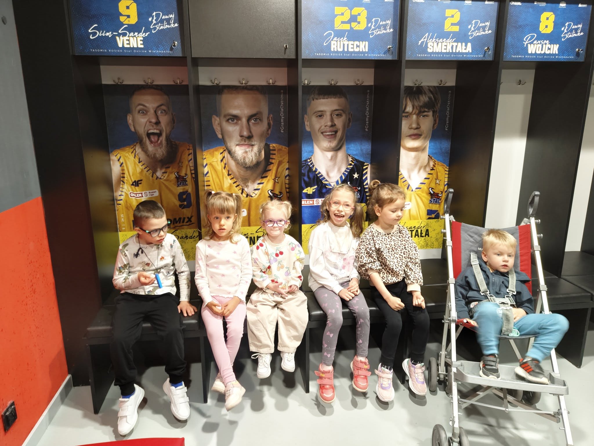 Sześcioro dzieci siedzących w szatni sportowców. Czworo siedzi na ławce, jeden chłopiec we wózku. W tle zdjęcia koszykarzy.