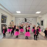 Dzieci ubrane na czarno, dziewczynki różowe spódniczki, chłopcy różowe krawaty stoją na szarym dywanie i tańczą w parach lub pojedynczo.