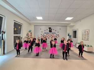 Dzieci ubrane na czarno, dziewczynki różowe spódniczki, chłopcy różowe krawaty stoją na szarym dywanie i tańczą w parach lub pojedynczo.