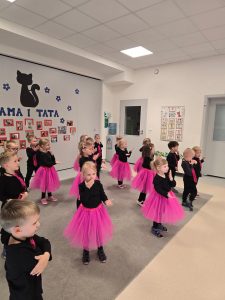 Dzieci ubrane na czarno, dziewczynki różowe spódniczki, chłopcy różowe krawaty stoją na szarym dywanie i tańczą w parach lub pojedynczo.