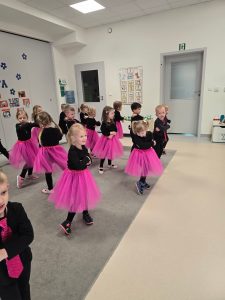 Dzieci ubrane na czarno, dziewczynki różowe spódniczki, chłopcy różowe krawaty stoją na szarym dywanie i tańczą w parach lub pojedynczo.