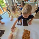 Dzieci siedzą przy białym stole i tworzą własną czekoladę z produktów, które znajdują się na stole.