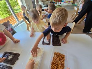 Dzieci siedzą przy białym stole i tworzą własną czekoladę z produktów, które znajdują się na stole.