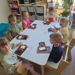 Dzieci siedzą przy białym stole i tworzą własną czekoladę z produktów, które znajdują się na stole.