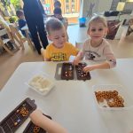 Dzieci siedzą przy białym stole i tworzą własną czekoladę z produktów, które znajdują się na stole.