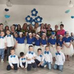 Grupa 24 dzieci wraz z 10 zaproszonymi gośćmi i 3 paniami wychowawczyniami pozuje do zdjęcia na tle dekoracji (kwiatów biało niebieskich). Wszyscy są elegancko ubrani i uśmiechają się.
