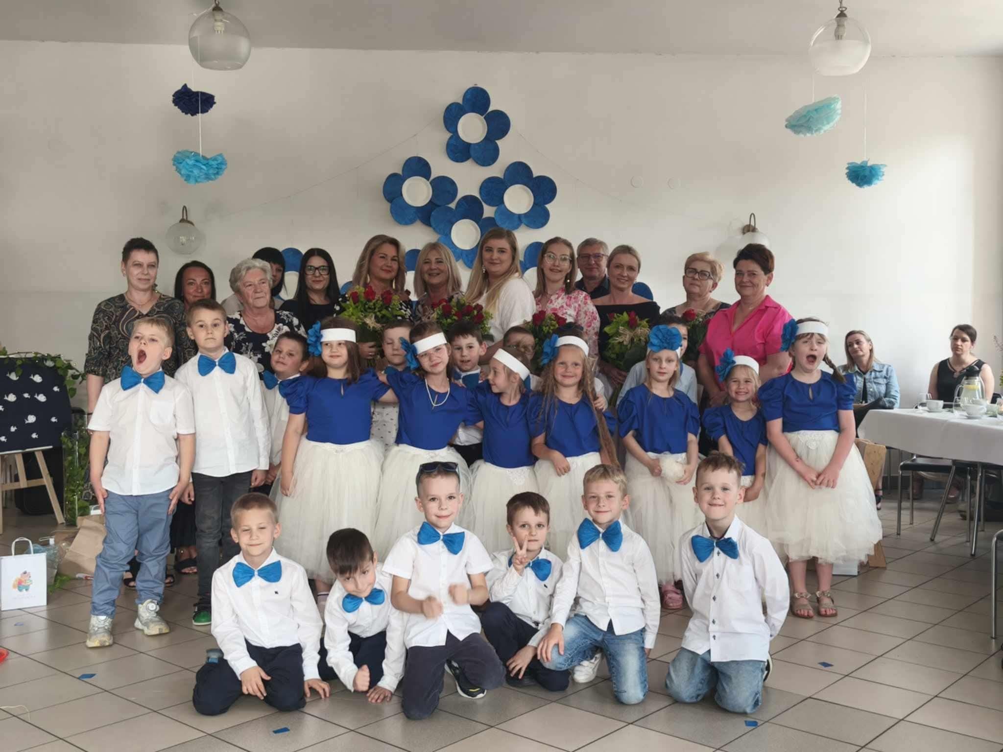 Grupa 24 dzieci wraz z 10 zaproszonymi gośćmi i 3 paniami wychowawczyniami pozuje do zdjęcia na tle dekoracji (kwiatów biało niebieskich). Wszyscy są elegancko ubrani i uśmiechają się.