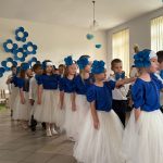 Grupa dzieci kończących przedszkole występuje na uroczystości dla swoich rodziców i zaproszonych gości. Dzieci są odświętnie ubrane, dziewczynki mają długie, tiulowe spódniczki, na głowach białe opaski z niebieskim kwiatem i modrakowe bluzeczki z krótkim rękawem. Chłopcy mają modrakowe muchy przy kołnierzykach swoich białych koszul, ciemne długie spodnie. Wszystkie dzieci wyglądają elegancko. Tańczą parami, tańczą poloneza. Tak więc para za parą. W tle dekoracje.