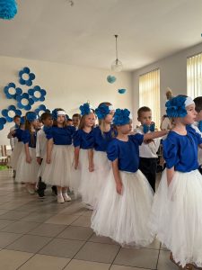 Grupa dzieci kończących przedszkole występuje na uroczystości dla swoich rodziców i zaproszonych gości. Dzieci są odświętnie ubrane, dziewczynki mają długie, tiulowe spódniczki, na głowach białe opaski z niebieskim kwiatem i modrakowe bluzeczki z krótkim rękawem. Chłopcy mają modrakowe muchy przy kołnierzykach swoich białych koszul, ciemne długie spodnie. Wszystkie dzieci wyglądają elegancko. Tańczą parami, tańczą poloneza. Tak więc para za parą. W tle dekoracje.