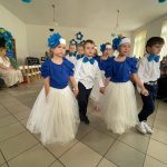 Grupa dzieci kończących przedszkole występuje na uroczystości dla swoich rodziców i zaproszonych gości. Dzieci są odświętnie ubrane, dziewczynki mają długie, tiulowe spódniczki, na głowach białe opaski z niebieskim kwiatem i modrakowe bluzeczki z krótkim rękawem. Chłopcy mają modrakowe muchy przy kołnierzykach swoich białych koszul, ciemne długie spodnie. Wszystkie dzieci wyglądają elegancko. Tańczą parami, tańczą poloneza. Tak więc para za parą. W tle dekoracje.