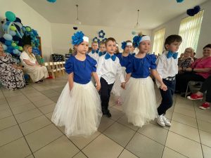 Grupa dzieci kończących przedszkole występuje na uroczystości dla swoich rodziców i zaproszonych gości. Dzieci są odświętnie ubrane, dziewczynki mają długie, tiulowe spódniczki, na głowach białe opaski z niebieskim kwiatem i modrakowe bluzeczki z krótkim rękawem. Chłopcy mają modrakowe muchy przy kołnierzykach swoich białych koszul, ciemne długie spodnie. Wszystkie dzieci wyglądają elegancko. Tańczą parami, tańczą poloneza. Tak więc para za parą. W tle dekoracje.