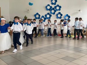 Grupa dzieci kończących przedszkole występuje na uroczystości dla swoich rodziców i zaproszonych gości. Dzieci są odświętnie ubrane, dziewczynki mają długie, tiulowe spódniczki, na głowach białe opaski z niebieskim kwiatem i modrakowe bluzeczki z krótkim rękawem. Chłopcy mają modrakowe muchy przy kołnierzykach swoich białych koszul, ciemne długie spodnie. Wszystkie dzieci wyglądają elegancko. Tańczą parami, tańczą poloneza. Tak więc para za parą. W tle dekoracje.