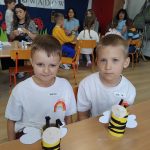 Dwóch chłopców w białych koszulkach z napisem "Przedszkole nr 12 Tęcza" z przodu i z numerem 12 z tyłu koszulki. Chłopcy pozują do zdjęcia podczas konkursu ekologicznego. Uśmiechają się, w tle widać inne dzieci - pozostałych uczestników konkursu.