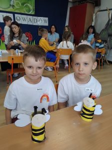 Dwóch chłopców w białych koszulkach z napisem "Przedszkole nr 12 Tęcza" z przodu i z numerem 12 z tyłu koszulki. Chłopcy pozują do zdjęcia podczas konkursu ekologicznego. Uśmiechają się, w tle widać inne dzieci - pozostałych uczestników konkursu.
