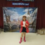 Dziewczynka w okularach, białej bluzce, krótkich czarnych spodenkach, rajstopach w serduszka i kaloszach na nogach. Dziecko stoi na tle fototapety przedstawiającej Big Ben w Londynie. W rękach trzyma prezent i dyplom za udział w konkursie piosenki angielskiej.