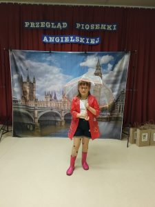 Dziewczynka w okularach, białej bluzce, krótkich czarnych spodenkach, rajstopach w serduszka i kaloszach na nogach. Dziecko stoi na tle fototapety przedstawiającej Big Ben w Londynie. W rękach trzyma prezent i dyplom za udział w konkursie piosenki angielskiej.