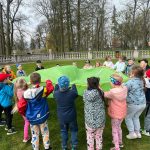 Grupa 20 dzieci w wieku przedszkolnym bawi się chustą animacyjną w kolorze zielonym na terenie parku przy zamku w Gutowie. Wszyscy są weseli i uśmiechnięci.