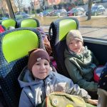 Dwoje dzieci (chłopiec i dziewczynka) siedzi w autobusie. Na głowach mają czapki zimowe. Chłopiec ma okulary. Obydwoje się uśmiechają, W tle widać parking z samochodami oraz budynek przedszkola nr 12.