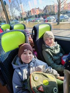 Dwoje dzieci (chłopiec i dziewczynka) siedzi w autobusie. Na głowach mają czapki zimowe. Chłopiec ma okulary. Obydwoje się uśmiechają, W tle widać parking z samochodami oraz budynek przedszkola nr 12.