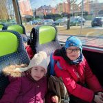 Dwoje dzieci (chłopiec i dziewczynka) siedzi w autobusie. Na głowach mają czapki zimowe. Chłopiec ma okulary. Obydwoje się uśmiechają, W tle widać parking z samochodami oraz budynek przedszkola nr 12.