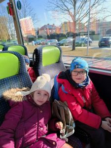 Dwoje dzieci (chłopiec i dziewczynka) siedzi w autobusie. Na głowach mają czapki zimowe. Chłopiec ma okulary. Obydwoje się uśmiechają, W tle widać parking z samochodami oraz budynek przedszkola nr 12.