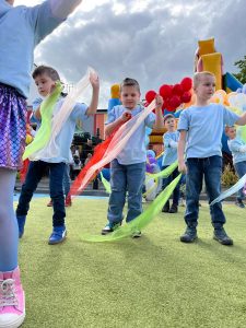 Dzieci ubrane w błękitne koszulki z krótkim rękawem i spódniczki w "rybią łuskę" tańczą trzymając w dłoniach kolorowe chusty. W tle widać budynek przedszkola nr 12.