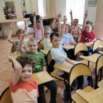 Dzieci siedzą na krzesłach w sali i zgłaszają się do odpowiedzi