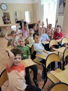 Dzieci siedzą na krzesłach w sali i zgłaszają się do odpowiedzi
