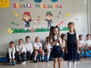 Dwie dziewczynki stoją i recytują wiersz, reszta dzieci siedzi w tyle na lawkach