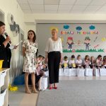 Dzieci siedzą na ławkach, dwie nauczycielki stoją i słuchają przemowy rodzica