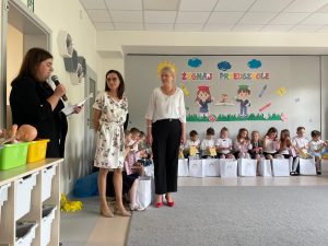 Dzieci siedzą na ławkach, dwie nauczycielki stoją i słuchają przemowy rodzica
