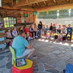 Dzieci siedzą w półkolu na ławkach w altanie i słuchają Pana opowiadającego o zwierzętach