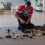 Mężczyzna ubrany w koszulkę klubu Ostrovia rozkłada na szarym dywanie swoje trofea sportowe.