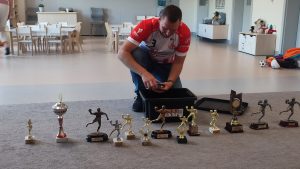 Mężczyzna ubrany w koszulkę klubu Ostrovia rozkłada na szarym dywanie swoje trofea sportowe.