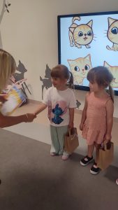 Grupka dzieci stoi obok siebie. Kobieta ubrana w czarną bluzkę wręcza dziecku dyplom.
