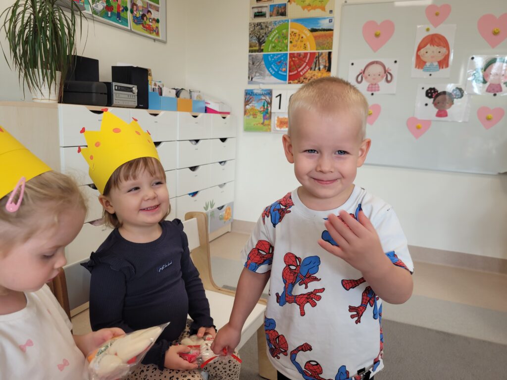 Dwie dziewczynki siedzą na krzesłach. Chłopiec wręcza upominek.